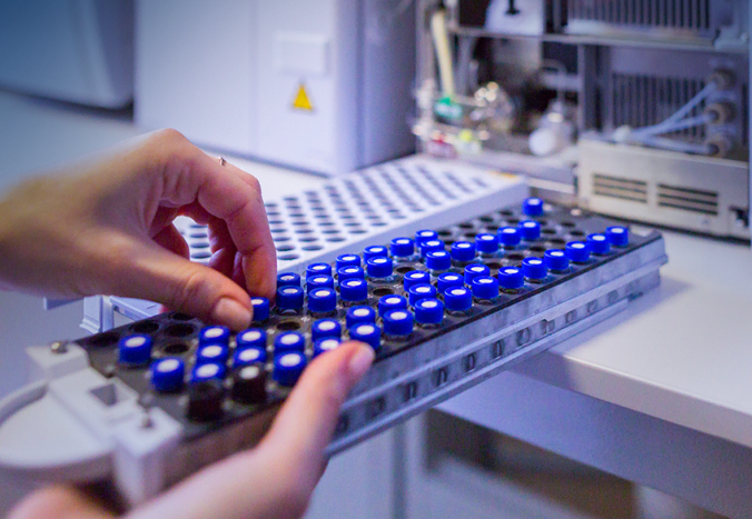 Les mains d’une personne placent ou retirent de petits flacons à bouchon bleu d’un plateau, préparant probablement des échantillons pour une analyse scientifique dans un laboratoire avec un équipement en arrière-plan.
