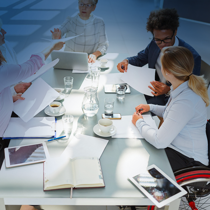Quatre professionnels sont assis autour d'une table, avec des papiers, des ordinateurs portables, des carnets et des tasses à café, en pleine discussion. Une femme est en fauteuil roulant. La lumière du jour entre par les fenêtres.