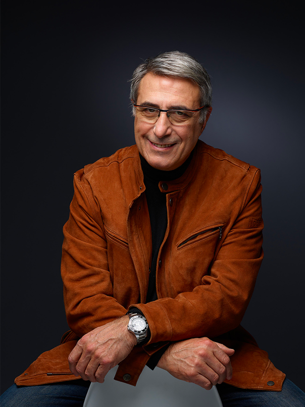 Un homme souriant aux cheveux gris et aux lunettes, portant une veste en daim marron et une chemise noire, est assis, les bras croisés sur un fond sombre.
