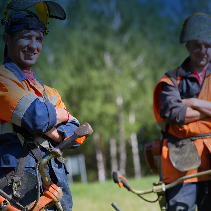 Deux ouvriers, vêtus de gilets de sécurité orange et de casques, se tiennent dehors, débroussailleuses à la main. Ils sourient et se tiennent dans une zone herbeuse avec des arbres en arrière-plan, sous un soleil radieux.