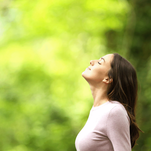 Une femme aux longs cheveux bruns se tient à l'extérieur, les yeux fermés et la tête légèrement inclinée vers le haut, respirant paisiblement l'air frais sur un fond vert flou.