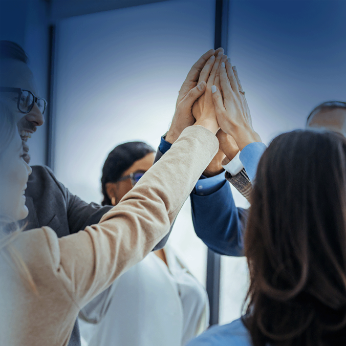 Un groupe de professionnels debout en cercle, joignant les mains dans un geste de high-five, souriant et célébrant le travail d'équipe dans un environnement de bureau moderne.