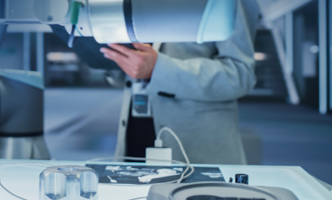 Une personne vêtue d'un costume de couleur claire utilise des machines de pointe ou un bras robotisé sur un poste de travail de haute technologie, avec divers appareils et composants électroniques visibles sur la table bien éclairée.