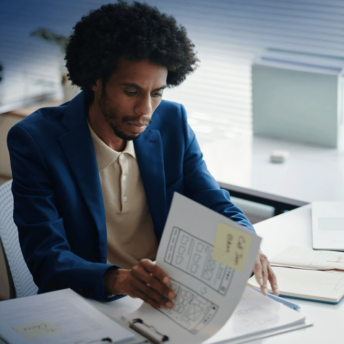 Un homme vêtu d'un blazer bleu examine les croquis de structures filaires d'un site Web à un bureau, entouré de documents et de dossiers dans un environnement de bureau moderne.