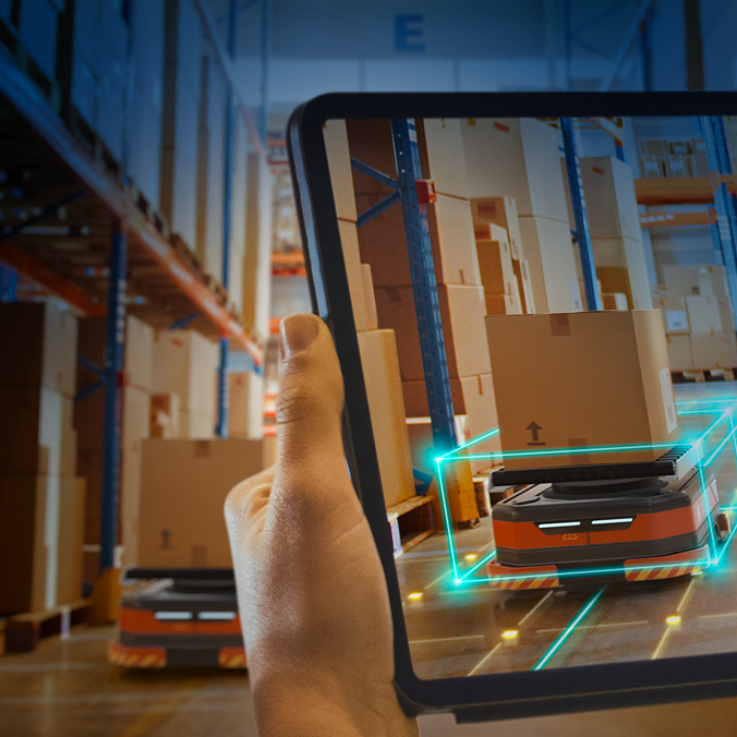 Une main tient une tablette montrant des visuels en réalité augmentée d'un véhicule robotisé transportant des cartons dans un entrepôt rempli d'étagères et de colis empilés.