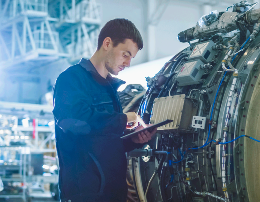 Un homme en uniforme de travail bleu utilise une tablette pendant qu'il inspecte ou travaille sur une grande machine complexe dans un environnement industriel ou de laboratoire.