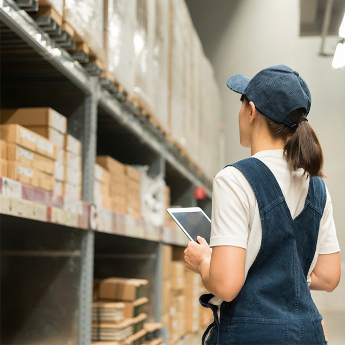 Un ouvrier en salopette et casquette tient une tablette tout en inspectant des étagères remplies de boîtes dans un entrepôt.