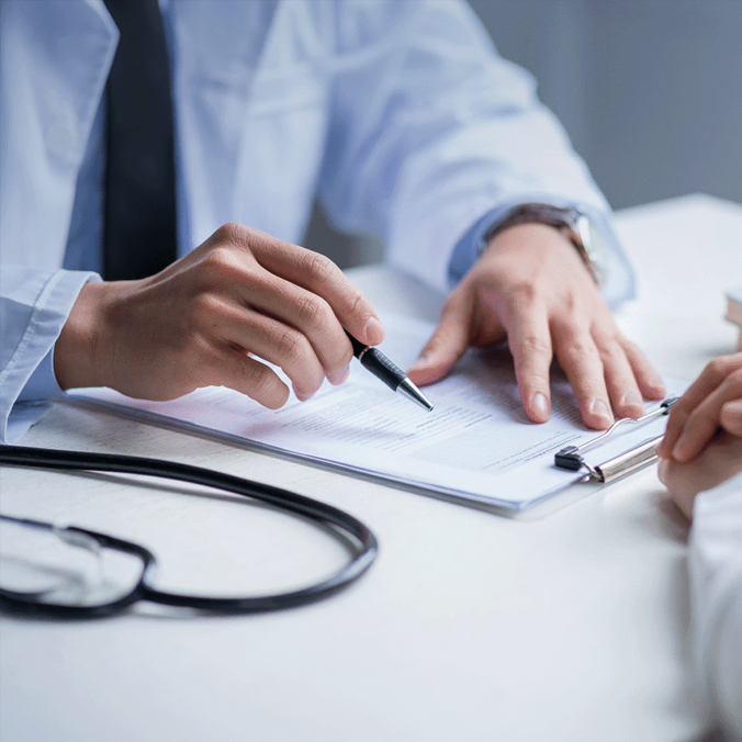 Un médecin en blouse blanche, un stylo à la main, discute de documents avec un patient. Un stéthoscope et des documents sont posés sur le bureau, suggérant une consultation médicale ou un bilan de santé.