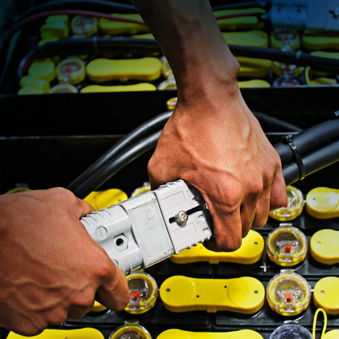 Gros plan de deux mains connectant un câble de charge à une batterie avec plusieurs cellules de batterie jaunes et connecteurs visibles en arrière-plan.
