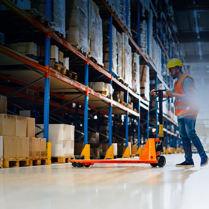 Un employé d'entrepôt, coiffé d'un casque jaune et d'un gilet de sécurité orange, manipule un transpalette et déplace des cartons sur de hautes étagères remplies de cartons et de colis. L'entrepôt spacieux est baigné d'une lumière vive.