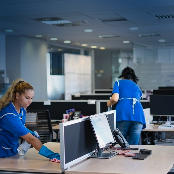 Deux femmes portant des uniformes bleus et des gants nettoient les bureaux et les écrans d'ordinateur dans un bureau moderne avec des chaises et des postes de travail vides, sous un éclairage fluorescent brillant.