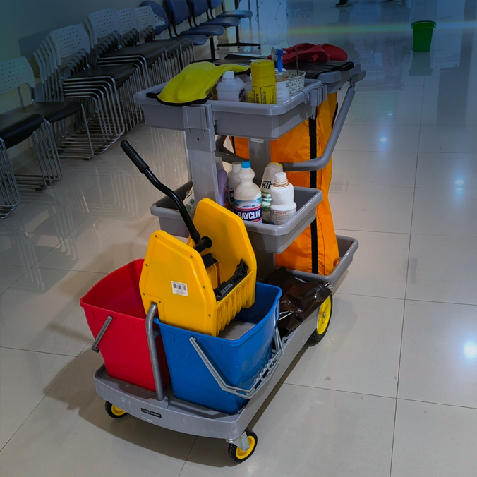 Un chariot de nettoyage de concierge avec un seau rouge et bleu, une serpillière, des produits de nettoyage et un panneau d'avertissement jaune se trouve sur un sol carrelé brillant près d'une rangée de chaises empilées dans un couloir.
