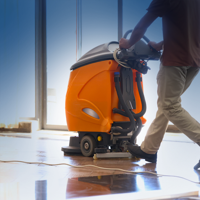 Une personne utilise une machine de nettoyage de sol industrielle orange sur une surface brillante et réfléchissante près de grandes fenêtres.