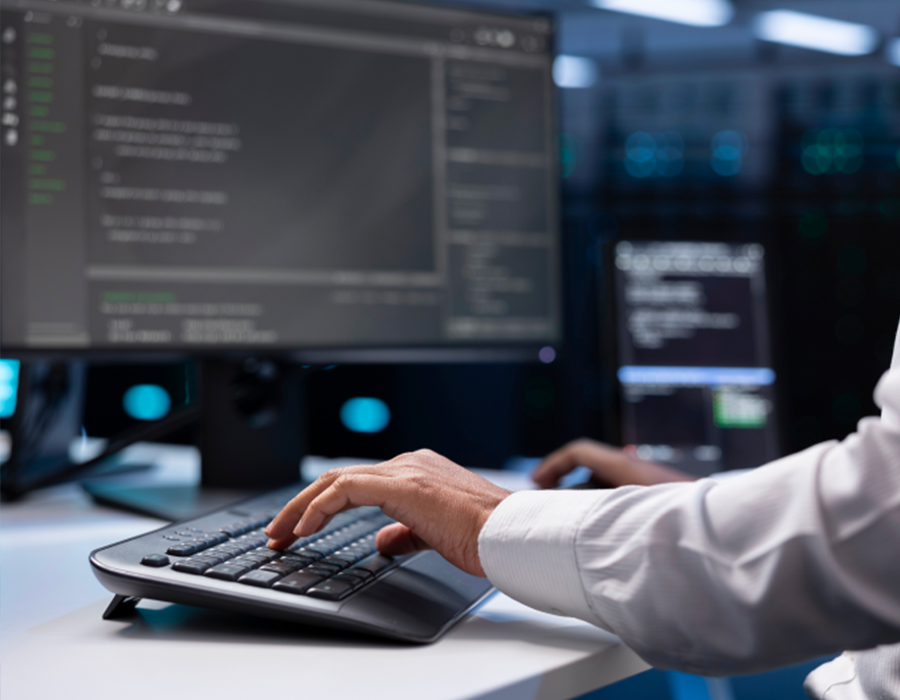 Une personne tape sur un clavier d'ordinateur tandis qu'un code s'affiche sur un écran dans un bureau moderne faiblement éclairé ou dans une salle de serveurs.