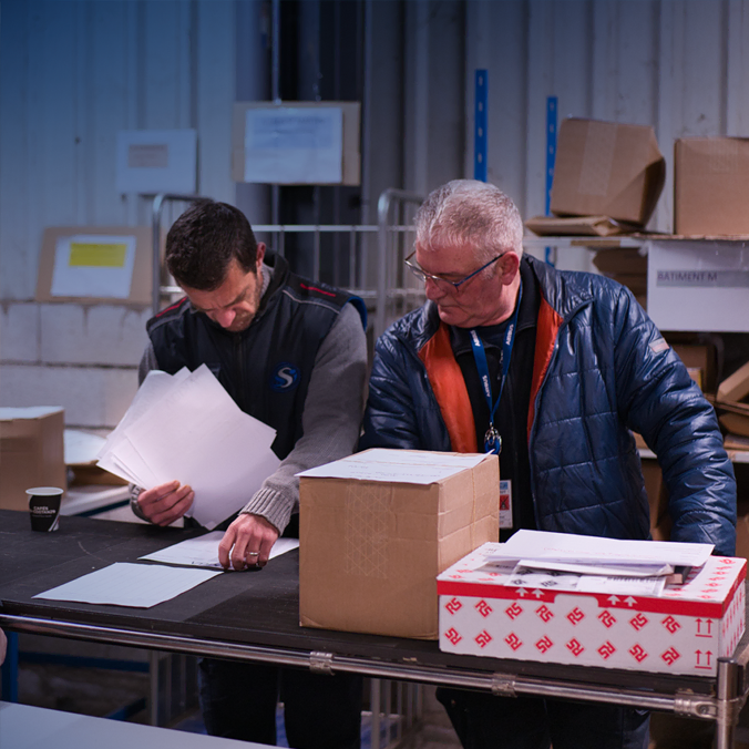 Deux hommes se tiennent à une table dans un entrepôt et examinent des documents et des cartons. L'un tient des papiers tandis que l'autre les examine. Des étagères et des colis sont visibles en arrière-plan.