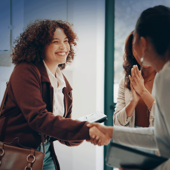 Une femme aux cheveux bouclés sourit en serrant la main d'une autre personne à l'intérieur. Deux autres personnes se tiennent à proximité, applaudissant et paraissant heureuses, suggérant un moment positif ou de félicitations.