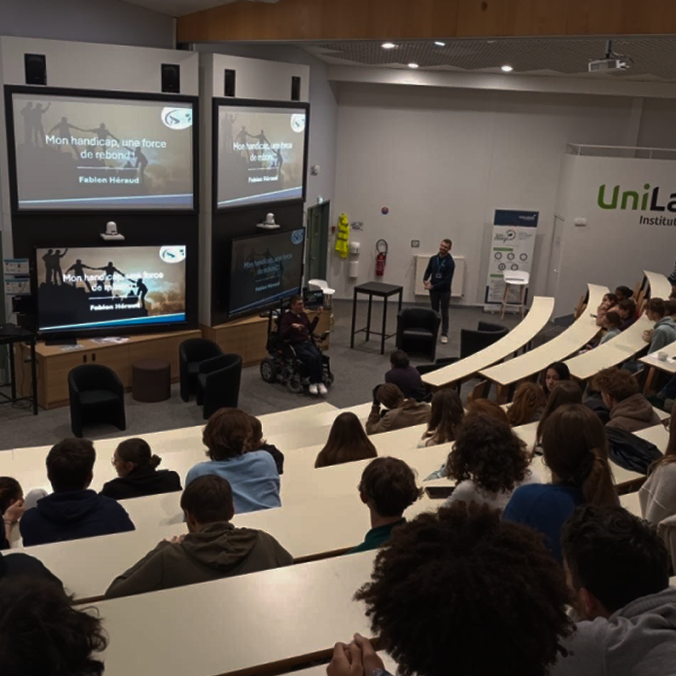 Un conférencier s'adresse à un public dans une salle de conférence avec des gradins. Plusieurs écrans diffusent une diapositive intitulée « Mon handicap, une force de réussite » de Fabien Hérout. Certains participants utilisent des ordinateurs portables ; une personne est en fauteuil roulant.