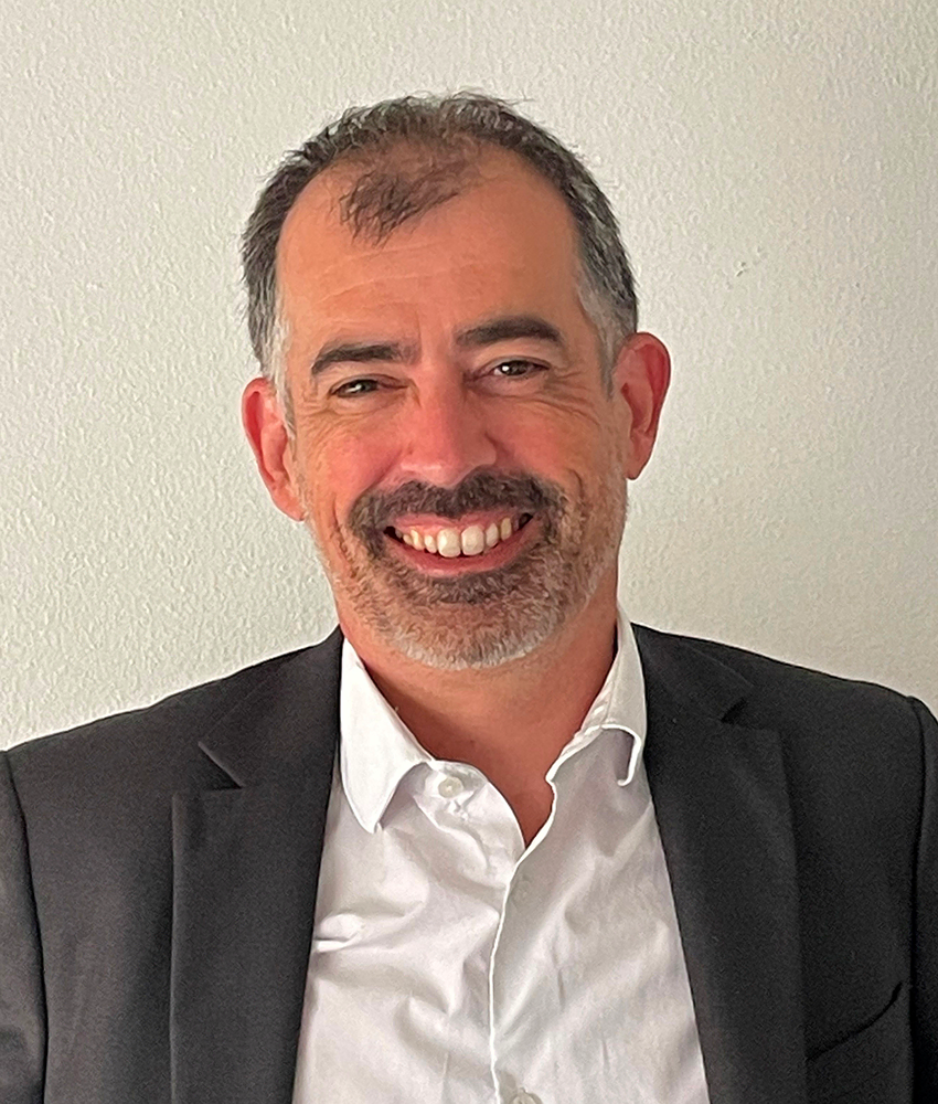 Un homme d'âge moyen aux cheveux bruns courts et à la barbe grise, vêtu d'un blazer noir et d'une chemise blanche, souriant devant un mur de couleur claire, se tient à côté de Priscilla DANJOU.