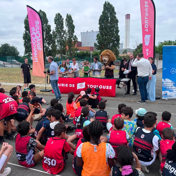 Un groupe d'enfants vêtus de maillots de sport est assis à l'extérieur, face à une scène où se trouvent des adultes, une mascotte de lion et des bannières de la mairie de Toulouse, suggérant un événement sportif ou une cérémonie de remise de prix.