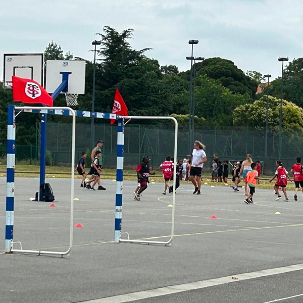 Des enfants et des adultes jouent au basket-ball en plein air sur un terrain, avec des drapeaux rouges sur les poteaux de but, des arbres et une clôture à l'arrière-plan. Certains joueurs portent des maillots rouges et des cônes délimitent l'aire de jeu.