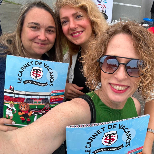 Trois femmes souriantes posent en plein air, tenant des livrets colorés intitulés Le Carnet de Vacances des Petits Stadistes avec des images de sport en bande dessinée sur les couvertures. Une femme porte des lunettes de soleil et tient l'appareil photo pour un selfie.