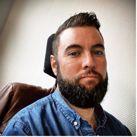 Un homme aux cheveux bruns courts et à la barbe fournie est assis sur une chaise brune, vêtu d'une chemise en jean bleu. Il regarde la caméra avec une expression neutre. L'arrière-plan est un mur blanc texturé.