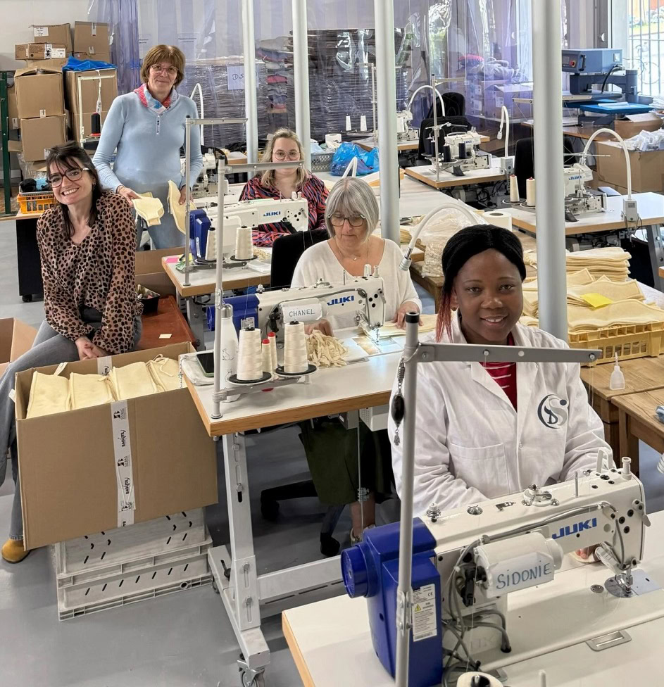 Cinq femmes sont assises devant des machines à coudre dans un atelier lumineux et sourient à la caméra. Des tissus et du matériel de couture sont posés sur les tables, et des étagères contenant du matériel sont visibles à l'arrière-plan.