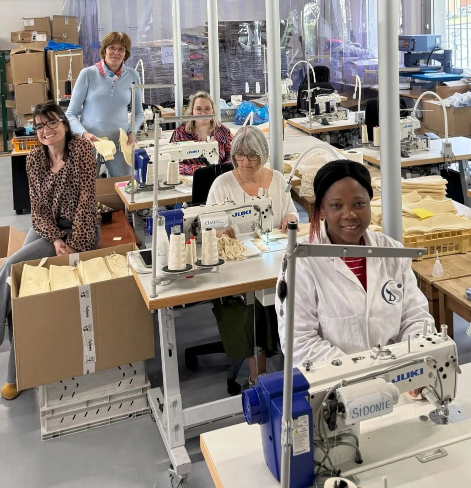 Cinq femmes sont assises devant des machines à coudre dans un atelier lumineux et sourient à la caméra. Des tissus et du matériel de couture sont posés sur les tables, et des étagères contenant du matériel sont visibles à l'arrière-plan.