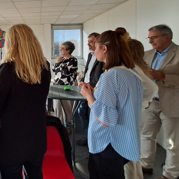 Un groupe de personnes se tient debout et discute autour d'une grande table ronde dans un intérieur lumineux, certains tenant des boissons, avec une fenêtre et des œuvres d'art colorées en arrière-plan.