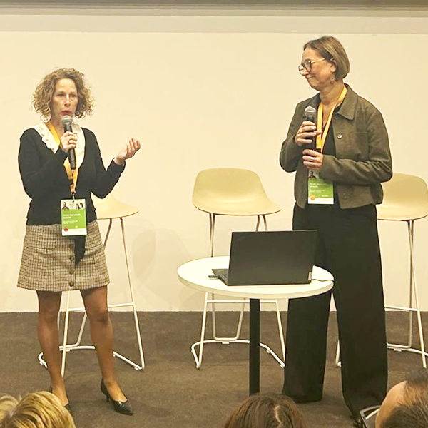 Deux femmes se tiennent sur une scène et parlent dans des microphones. L'une fait un geste de la main tandis que l'autre regarde. Derrière elles se trouvent des chaises vides et une table ronde avec un ordinateur portable. Un public est visible au premier plan.