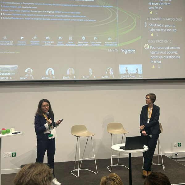 Deux femmes se tiennent sur une scène devant un grand écran affichant une réunion virtuelle avec des messages et les noms des participants. L'une des femmes parle avec un microphone, tandis que l'autre écoute. Deux chaises et une table se trouvent sur la scène.