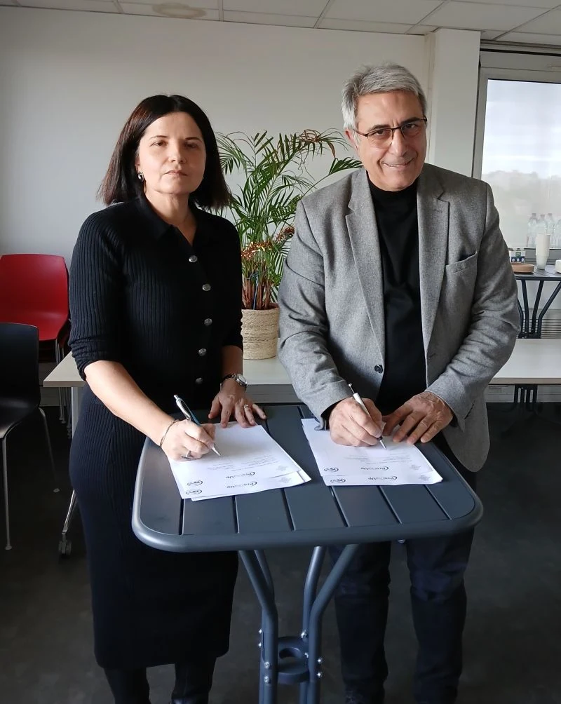 Une femme et un homme sont assis à une table, souriant et signant des documents. Ils sont à l'intérieur, avec des chaises, une plante et des rafraîchissements visibles à l'arrière-plan.