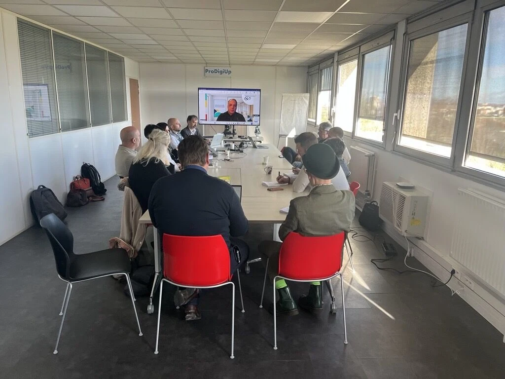 Dans un bureau moderne, un groupe de personnes est assis autour d'une table de conférence pour assister à une réunion, tandis qu'un participant se joint à la réunion par le biais d'un appel vidéo sur un grand écran situé au bout de la pièce.