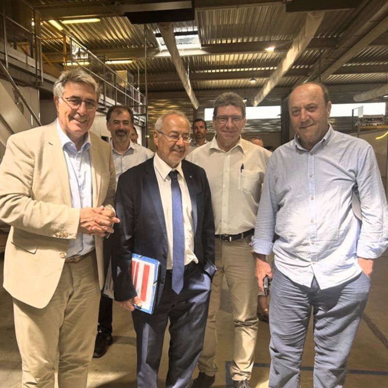 Cinq hommes se tiennent côte à côte dans un décor industriel ou semblable à un entrepôt, bien éclairé, et sourient à la caméra. L'un d'entre eux, en costume, tient un dossier, tandis que les autres portent une tenue professionnelle décontractée. On aperçoit au-dessus d'eux des poutres métalliques et des passerelles.