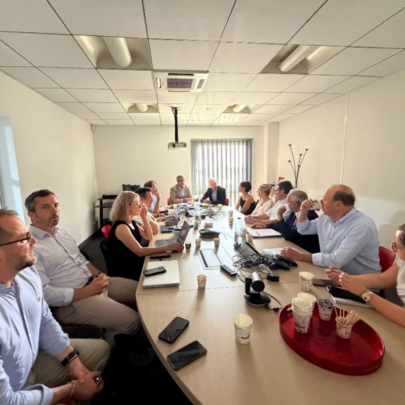 Un groupe de personnes est assis autour d'une grande table de conférence dans une salle de réunion lumineuse, en pleine discussion, avec devant elles des ordinateurs portables, des documents et des tasses de café. Un projecteur est suspendu au plafond.