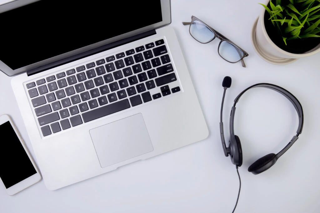 Un ordinateur portable, un smartphone, un casque avec micro, des lunettes et une plante en pot sont disposés sur un bureau blanc, vus d'en haut.
