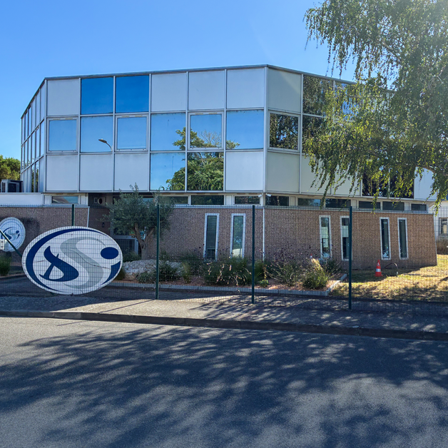 Un immeuble de bureaux moderne à deux étages, doté de grandes baies vitrées, entouré d'arbres et d'une clôture. Un grand panneau portant la lettre S est installé près de l'entrée. Le ciel est dégagé et ensoleillé.
