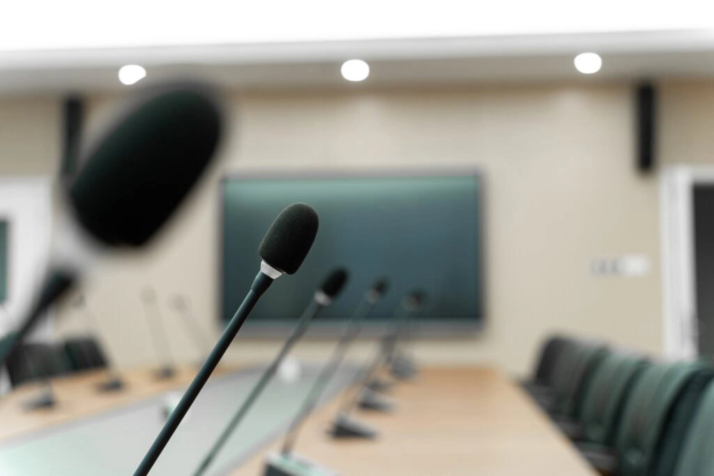 Gros plan sur des microphones posés sur une longue table de conférence dans une salle de réunion vide, avec des chaises disposées de part et d'autre et un grand écran en arrière-plan.