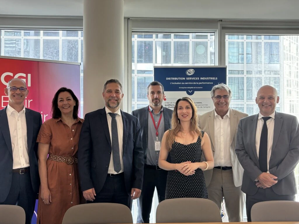 Sept personnes, quatre hommes et trois femmes, posent en souriant, vêtues de tenues professionnelles, dans une salle de réunion d'entreprise, avec de grandes fenêtres et deux bannières de l'entreprise en arrière-plan.